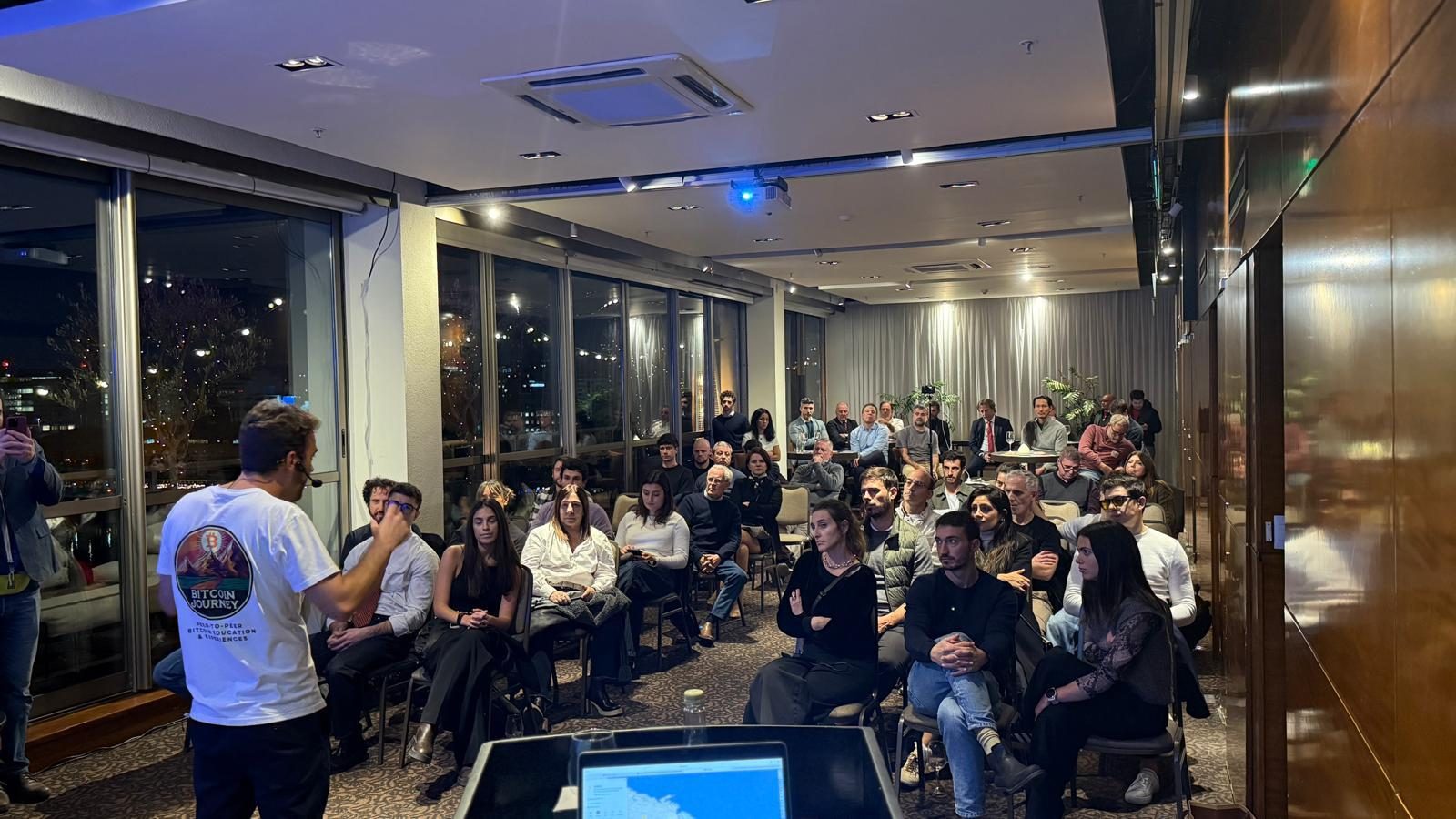 A Bitcoin Journey presenter facing a large seated audience at an evening session.