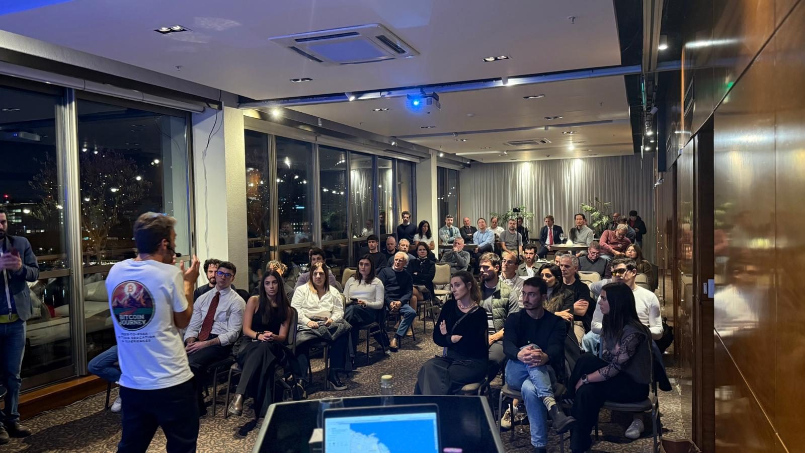 A Bitcoin Journey presenter speaking to about thirty seated people at an evening session, city lights visible through the windows behind them.