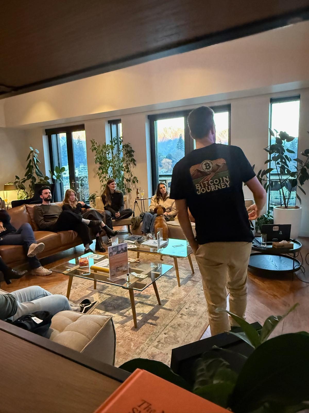 A Bitcoin Journey host talking with a small group seated on sofas in a lounge, mountains visible through the windows.