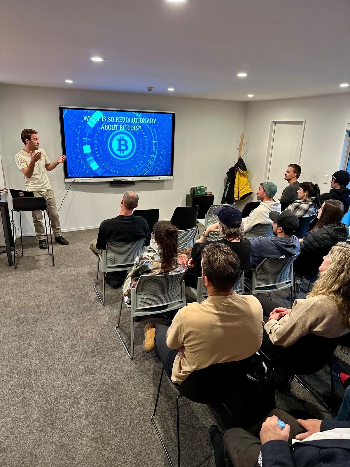 A presenter standing beside a screen that reads "What is so revolutionary about Bitcoin?" while a seated group watches.